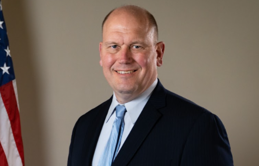 A man wearing a dark suit and light blue tie is smiling. 