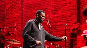 Rapper GZA in a black hoodie adjusts a mic stand against a very red brick wall.