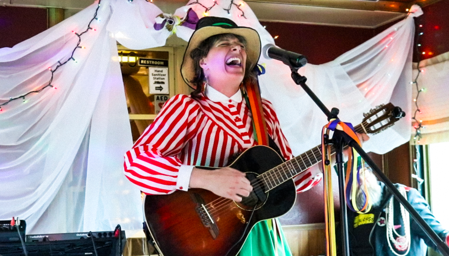 A woman wearing a red stripped shirt and a green skirt and hat plays guitar and sings in the dining car of a train. 