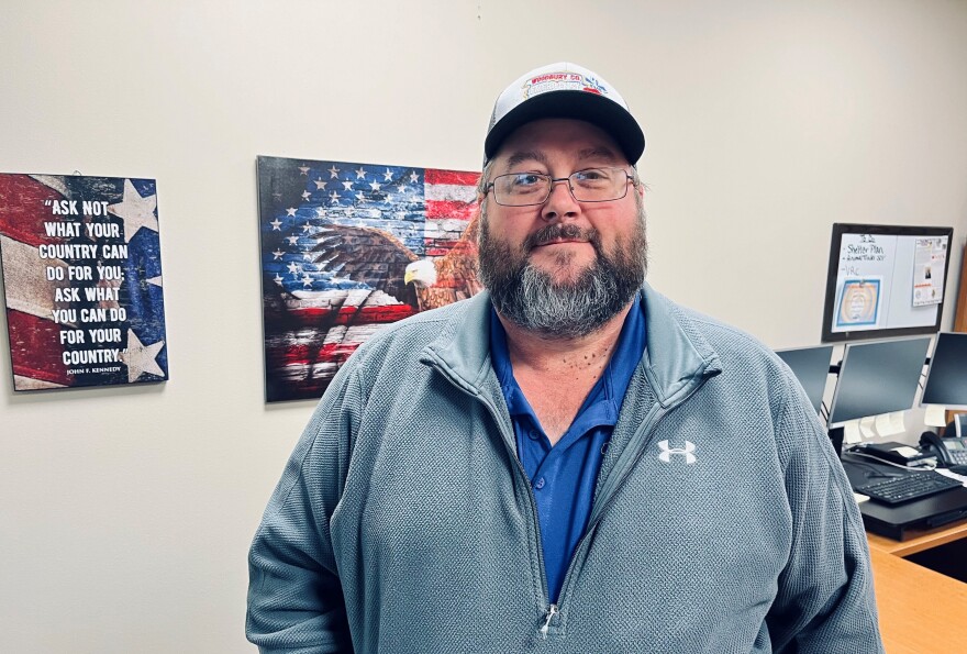 Man with a grey pullover and wearing a hat and glasses is standing in front of two patriotic pictures. They both have U.S. flags, one with an eagle and other other has the words "ask not what your country can do for you; ask what you can do for your country. Man also has full facial hair.