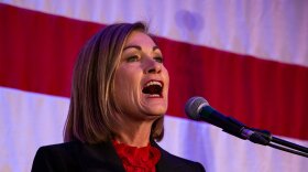 Gov. Kim Reynolds speaks into a microphone in front of an American flag. 