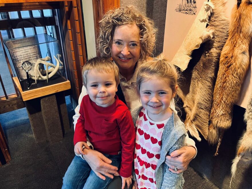 A smiling woman with curly blonde hair is holding a young boy who is wearing a burgundy top and a girl with a shirt with red and pink hearts. 