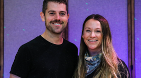 A man and woman dressed in black smile in front of a purple wall with laser dots.