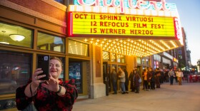 People in line outside a theater with a brightly lit, neon sign. A woman stands in front to take a photo of herself and the sign.