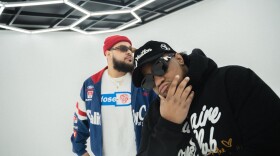 Two men in sunglasses, the one on the left wearing a blue sweatshirt and the one on the right wearing black, pose in an all white room with hexagonal light fixtures.