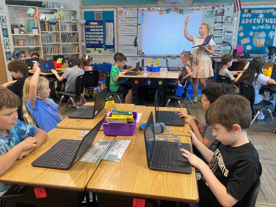 A teacher is in front of a class of 15 students who are sitting at tables in a classroom.