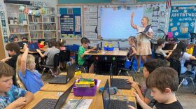 A teacher is in front of a class of 15 students who are sitting at tables in a classroom.