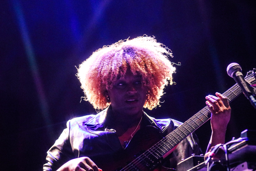 A curly blonde-haired woman in a leather jacket plays the guitar.