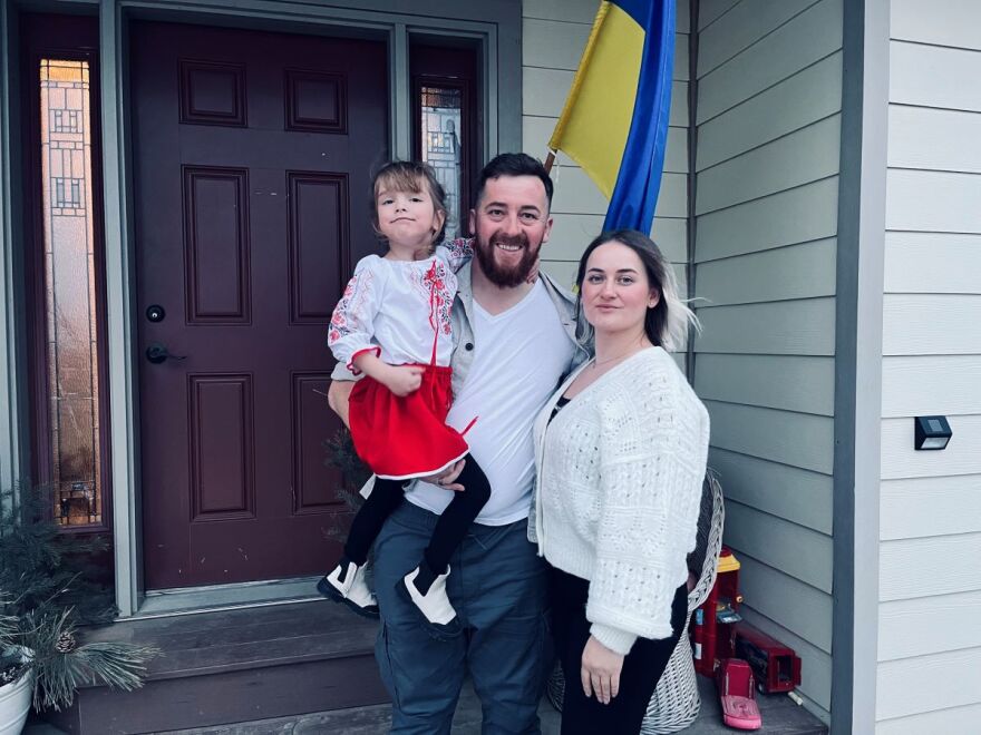 A man holds a little girl with a woman standing next to him. They are outside of a house with a Ukrainian flag behind them.