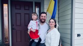 A man holds a little girl with a woman standing next to him. They are outside of a house with a Ukrainian flag behind them.