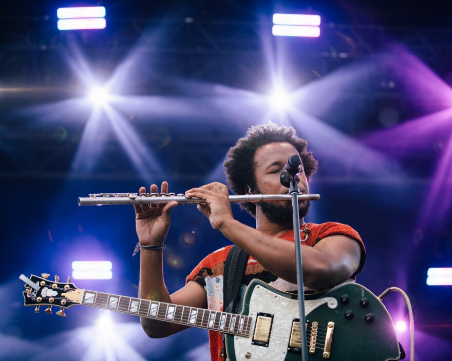  Cautious Clay plays the flute with a guitar around his person. Lights in the background of the photo frame him with purple and blue light. 