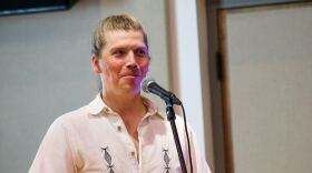 River Glen, a blonde man with his hair in a french braid, wears a white collared button up shirt in front of a khaki backdrop. 