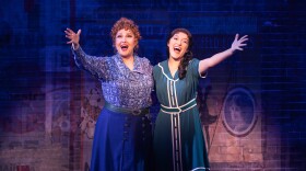 Two women sing on stage with their hands outstretched towards the audience.