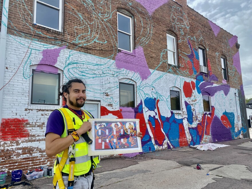 David Manzanares stands in front of his half finished mural in Sioux City. He holds the planned finished design.