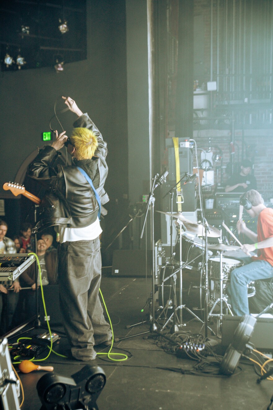 A man with a guitar around his neck screaming into a crowd stands next to his drummer. A sound tech can be seen in the background of the image. The photo is taken side stage at a rock concert. 