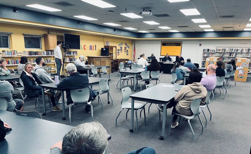 Several people are sitting in a school library for a school board meeting. They are sitting around tables with the school board sitting at the front.