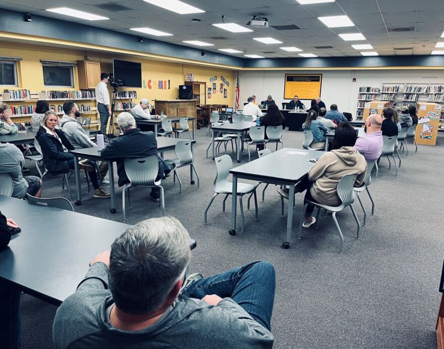 Several people are sitting in a school library for a school board meeting. They are sitting around tables with the school board sitting at the front.