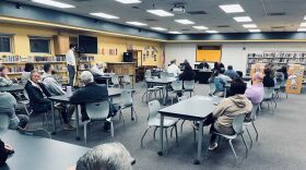 Several people are sitting in a school library for a school board meeting. They are sitting around tables with the school board sitting at the front.