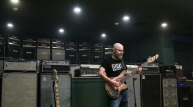 A bearded man with a bass guitar stands in front of a wall of over 80 amplifiers.