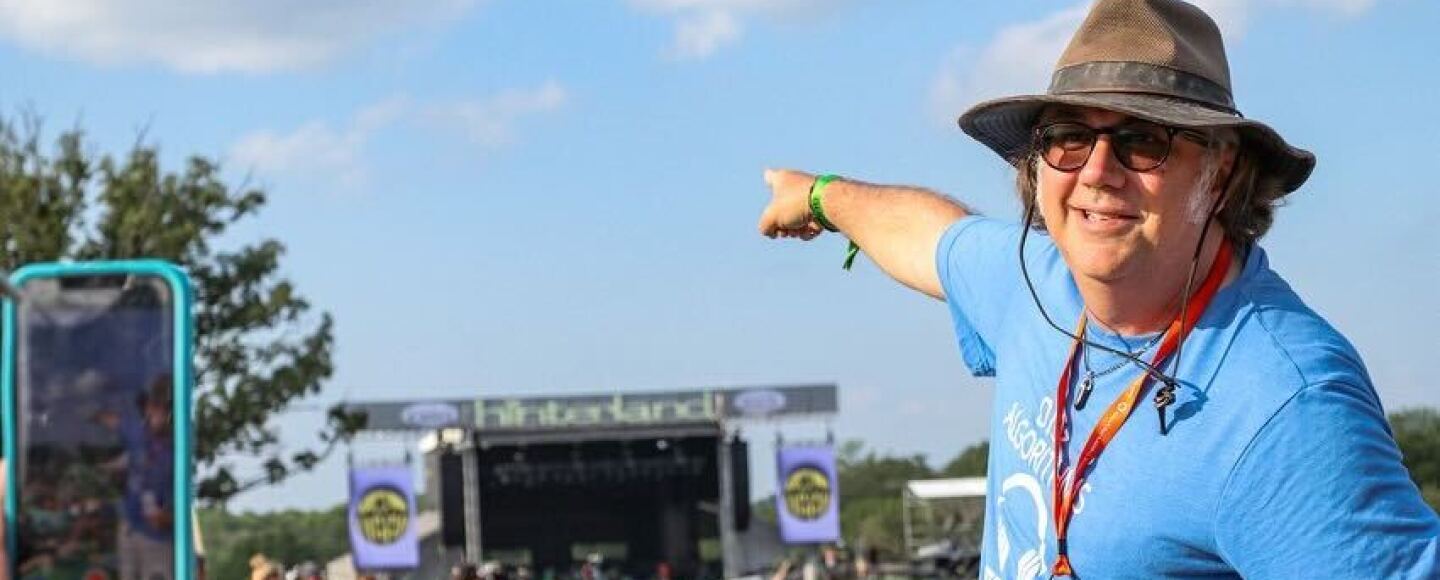 Tony Dehner wears an Indiana Jones style hat and a bright blue t-shirt and is standing outside in a field with a major music festival stage behind him.