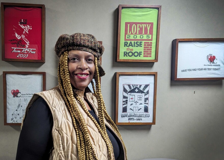 A woman stands facing the camera. On the wall behind her are several framed T-shirts with different messages of positivity.