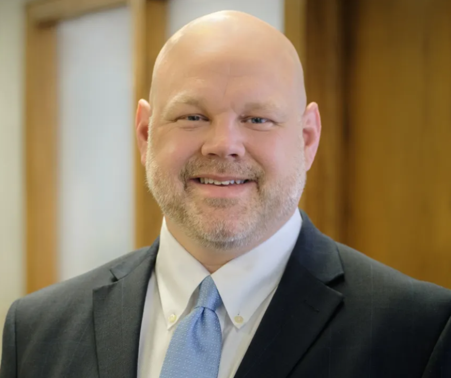 A bald man who is wearing a dark gray suit with a light blue tie is smiling.