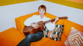 A horizontal photo of Abbie Sawyer holding an acoustic guitar, sitting relaxed on an orange corner couch. 