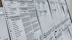 Sideways photo of sample ballots on a bulletin board. The ballots are white with black print and the word "sample" is in bigger print at the bottom of the paper.