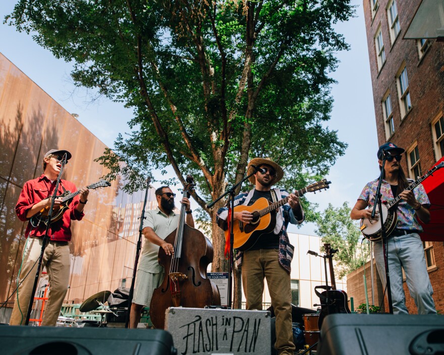 Four men stand on a stage with brick behind them playing stringed instruments.