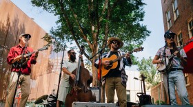 Four men stand on a stage with brick behind them playing stringed instruments.