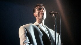 A still image of singer David Byrne from the 1983 film Stop Making Sense. Byrne is singing into a microphone on a stand in an oversized white suit, set against a dark background. 