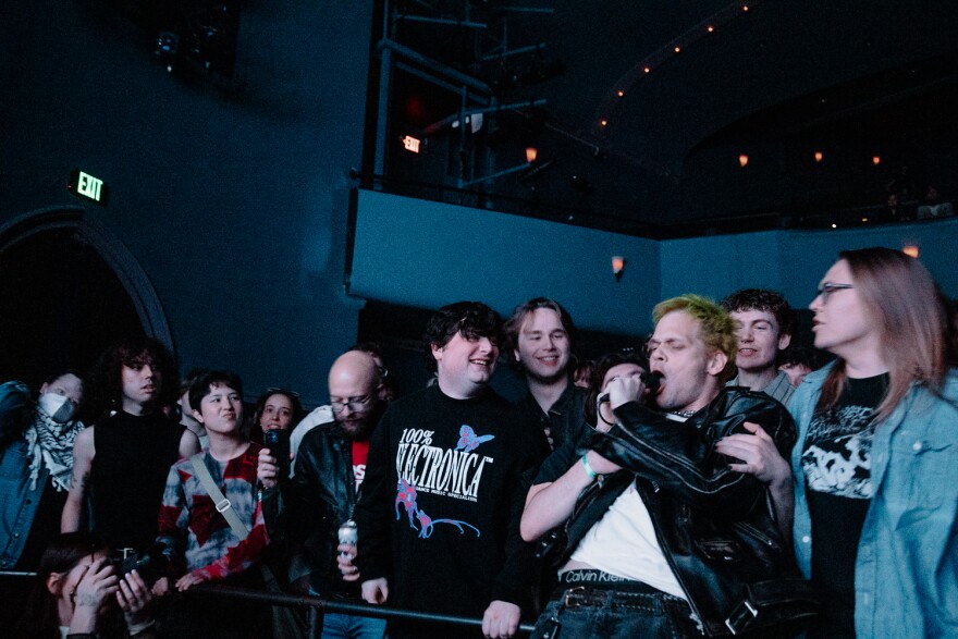 A man wearing a leather jacket with yellow hair sings into a microphone with his back to an audience directly behind him. 