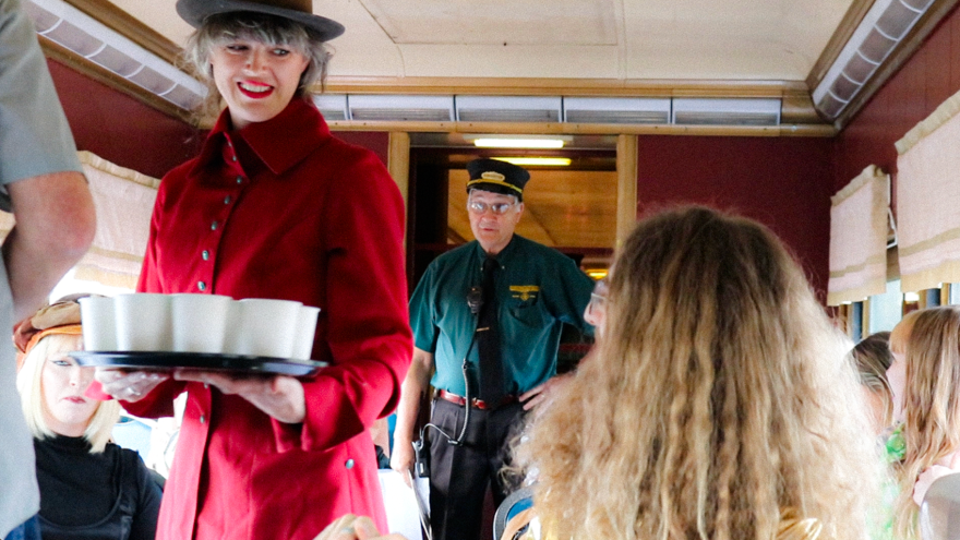 A woman in a red trench coat serves popcorn from small white cups and stands in the center of a train's dining car. Tables line the aisle, behind her the conductor stands. 