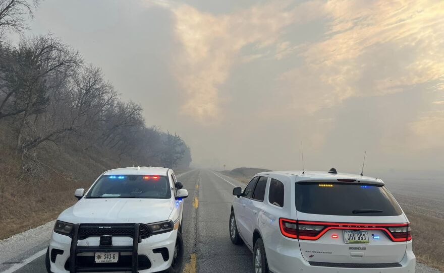 Two police cars sit in a street. Behind them, clouds of smoke rise up from the field fire.