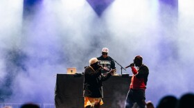  Big Boi and his band look at each other on stage at 80/35 in downtown Des Moines. There's smoke around them and purple lights as the crowd looks on in the foreground of the photo. 