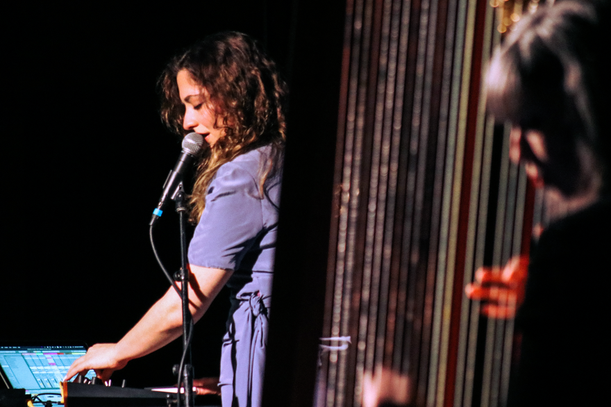 Two women perform, one at a switchboard singing into a microphone, the other plucking a harp.