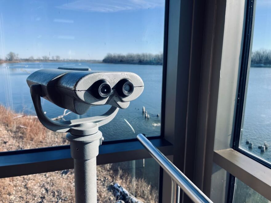 Photo shows a glass-covered observation area with a large grey binocular device with water in the background.