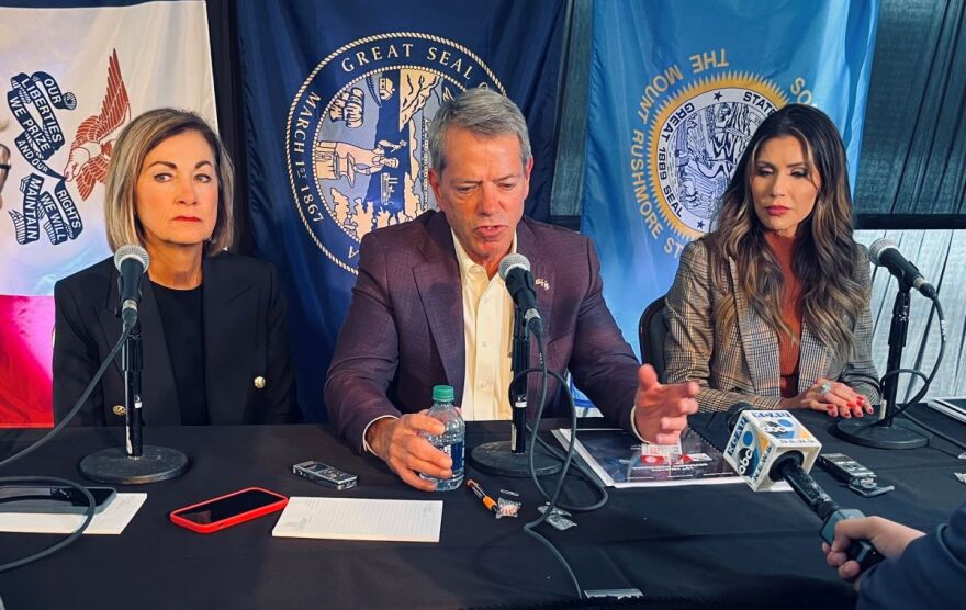 A man sits in front of a microphone with two women on each side. There are three state flags of Iowa, Nebraska, and South Dakota behind them. A reporter holds a microphone in front of the group.