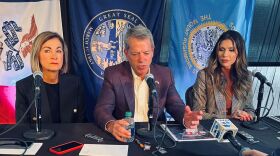 A man sits in front of a microphone with two women on each side. There are three state flags of Iowa, Nebraska, and South Dakota behind them. A reporter holds a microphone in front of the group.