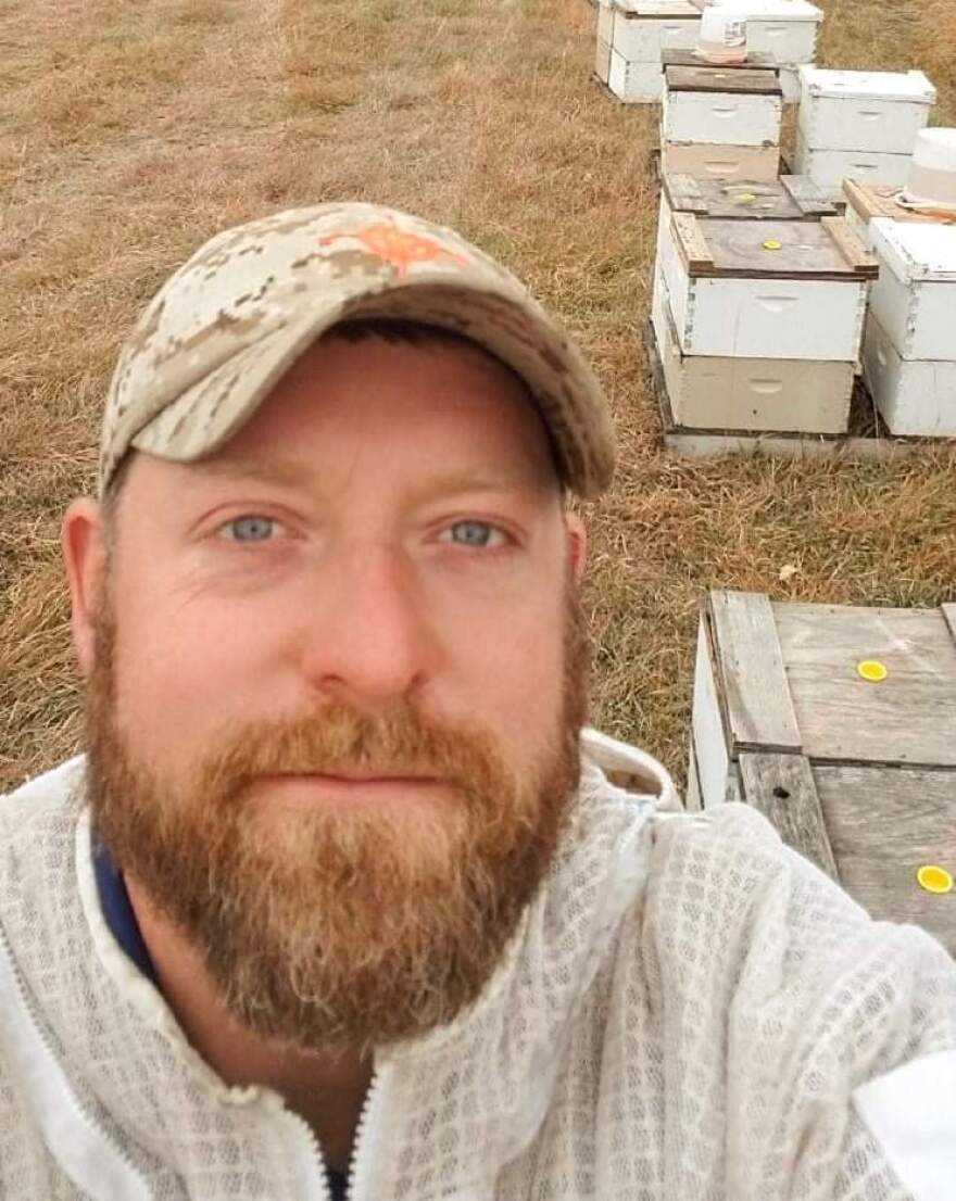 A man with a beard and a baseball cap stands in front of several white boxes holding bee colonies. 