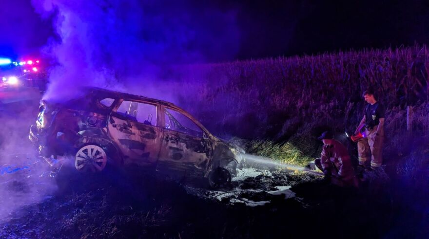 Firefighters use their hoses to extinguish a car fire in a ditch by a corn field during a dark night.