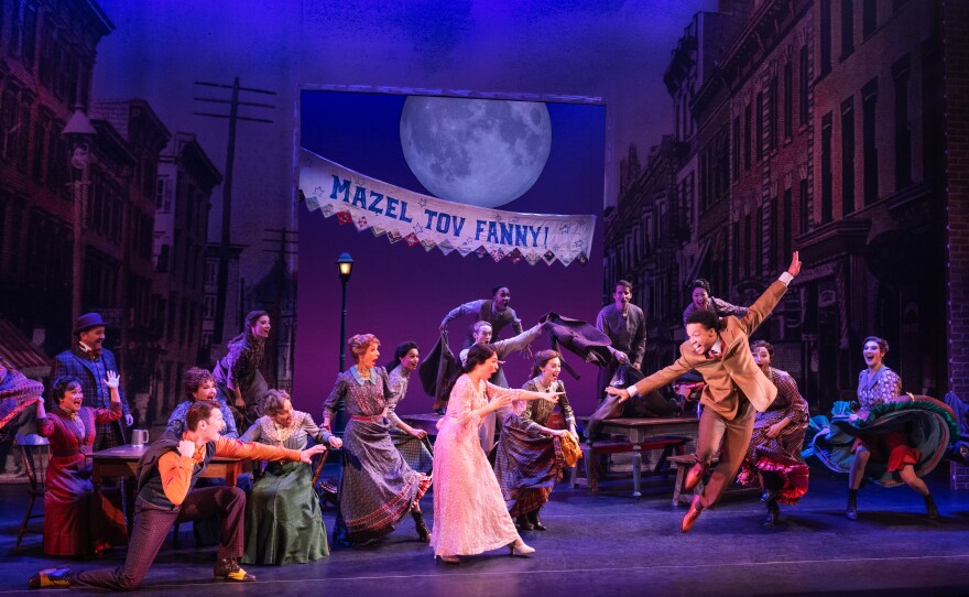 A group of actors dance on stage in front of a backdrop of a city street, which includes a banner that reads "Mazel Tov Fanny!"