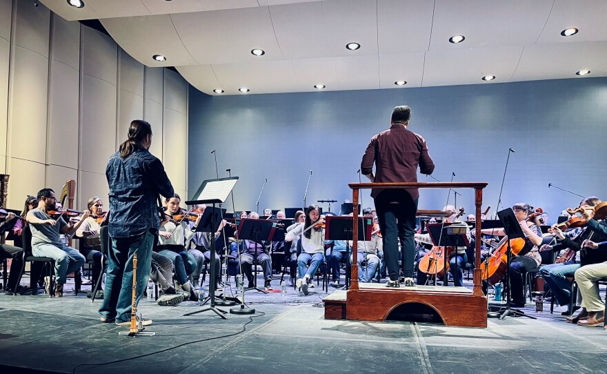 Image of a symphony rehearsal with an oboist standing to the left and the conductor in the center. They have their backs to camera. 