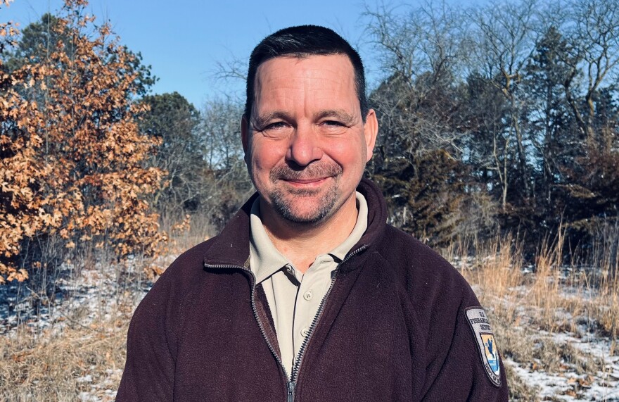 Man wearing a brown coat is smiling and standing in front of the woods.