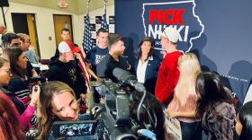 A crowd of about a dozen people stands around a woman with dark hair who is running for president. They are holding campaign signs and there are reporters with video cameras. 