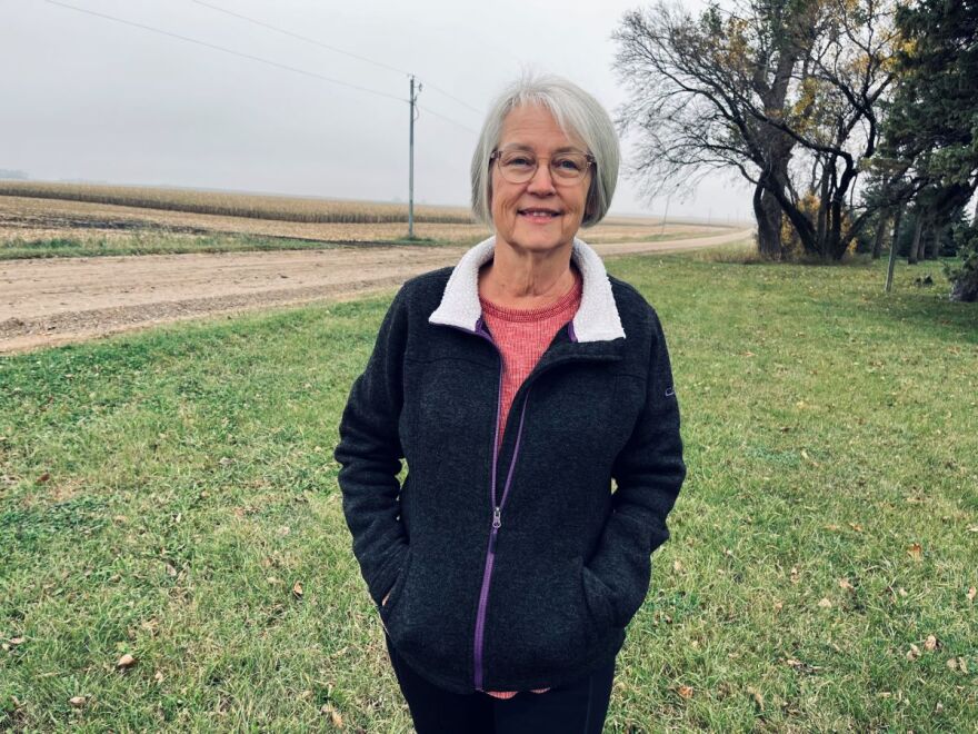 A smiling woman with grey hair and glasses is wearing a blue coat with a white color. She is standing in her lawn with a cornfield behind her.