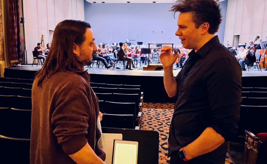 Two men are speaking with a group of orchestra performers sitting on a stage behind them.