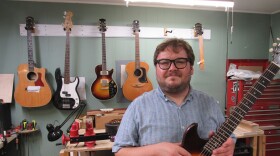 Dave Helmer stands in his guitar repair studio holding an electric sunburst guitar with a rack of tools and a workbench behind him. 