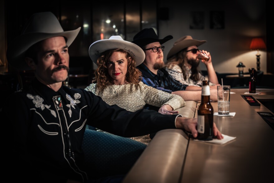 A color image of Katie and the Honky Tonks at a bar. Katie is facing the camera, while the others are looking over the bar.  