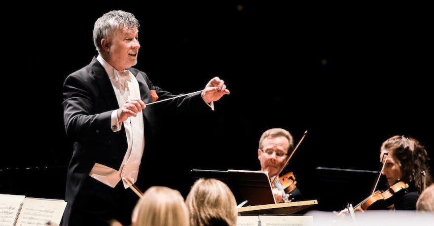 Conductor of a symphony orchestra holding a baton with outstretched arms over the musicians seated in front of him.